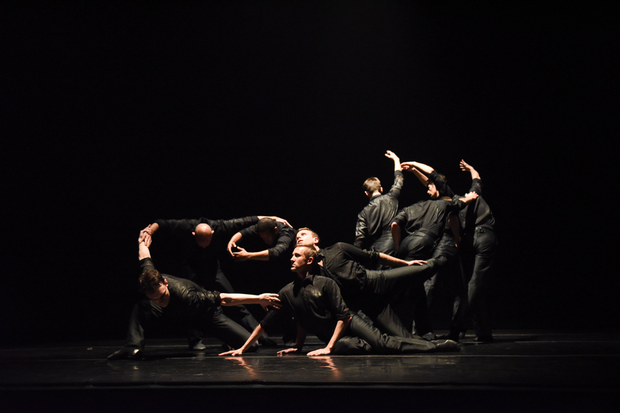 Men wearing all black curve their arms, some lunge to floor, and others bend their arms above their head with their backs facing the audience.